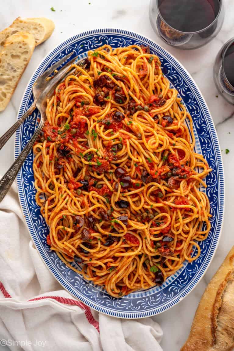 Overhead of platter of Pasta Puttanesca with two forks, glass of red wine and sliced baguette sitting beside