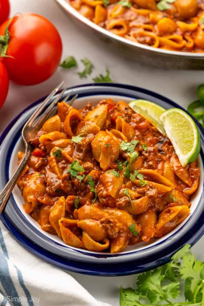 Bowl of One Pan Cheesy Taco Pasta garnished with two lime wedges and fresh cilantro and fork. Cilantro, tomatoes, and skillet sitting in background