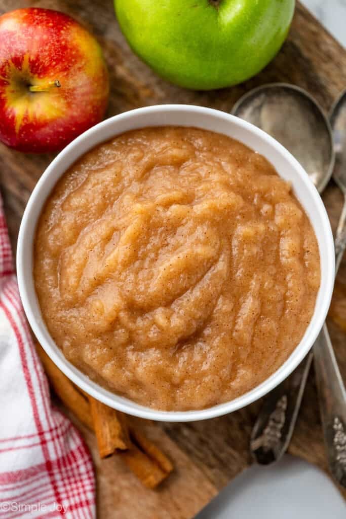 Overhead of white bowl of applesauce, with two apples, spoons, and cinnamon sticks surrounding bowl