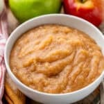 Bowl of homemade applesauce with two apples in background and cinnamon sticks sitting beside