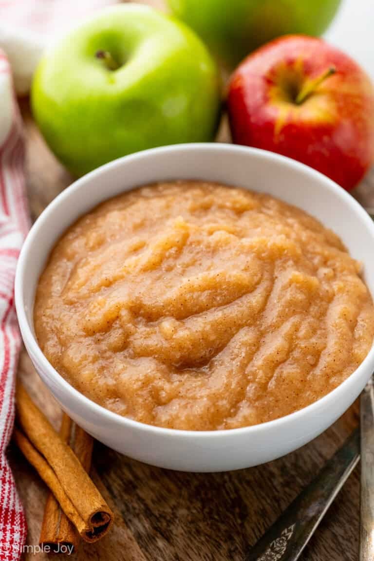 Bowl of homemade applesauce with two apples in background and cinnamon sticks sitting beside