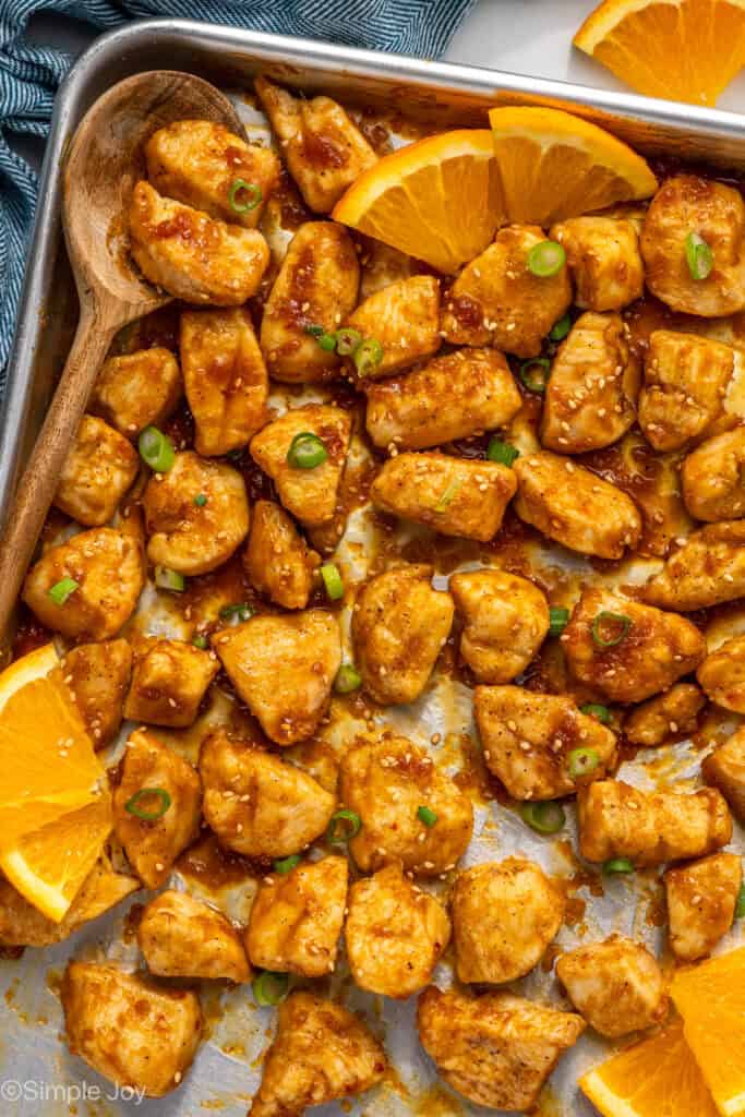 Overhead of baking sheet of Baked Orange Chicken with wooden spoon and orange slices