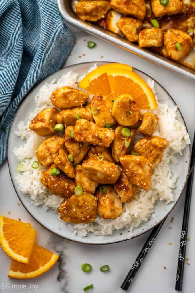 Overhead of plate of white rice topped with Baked Orange Chicken, garnished with orange slices and green onions. Chop sticks, orange slices, and baking sheet of orange chicken sitting beside