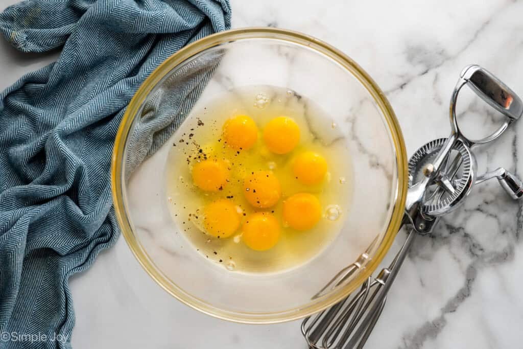 Overhead of glass bowl of eggs and seasonings with hand mixer beside to make breakfast burrito recipe