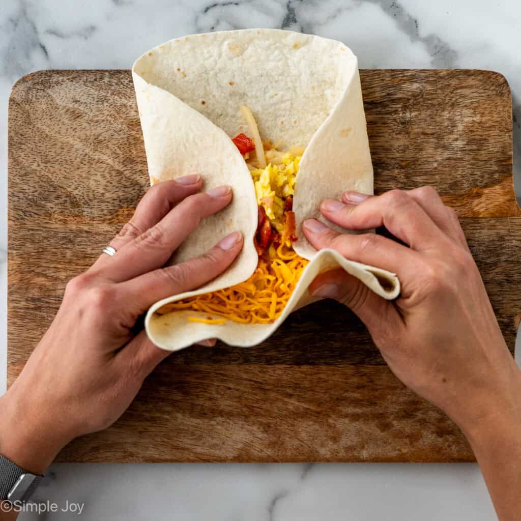 Overhead of woman's hands folding tortilla topped with Breakfast Burrito ingredients to make a breakfast burrito
