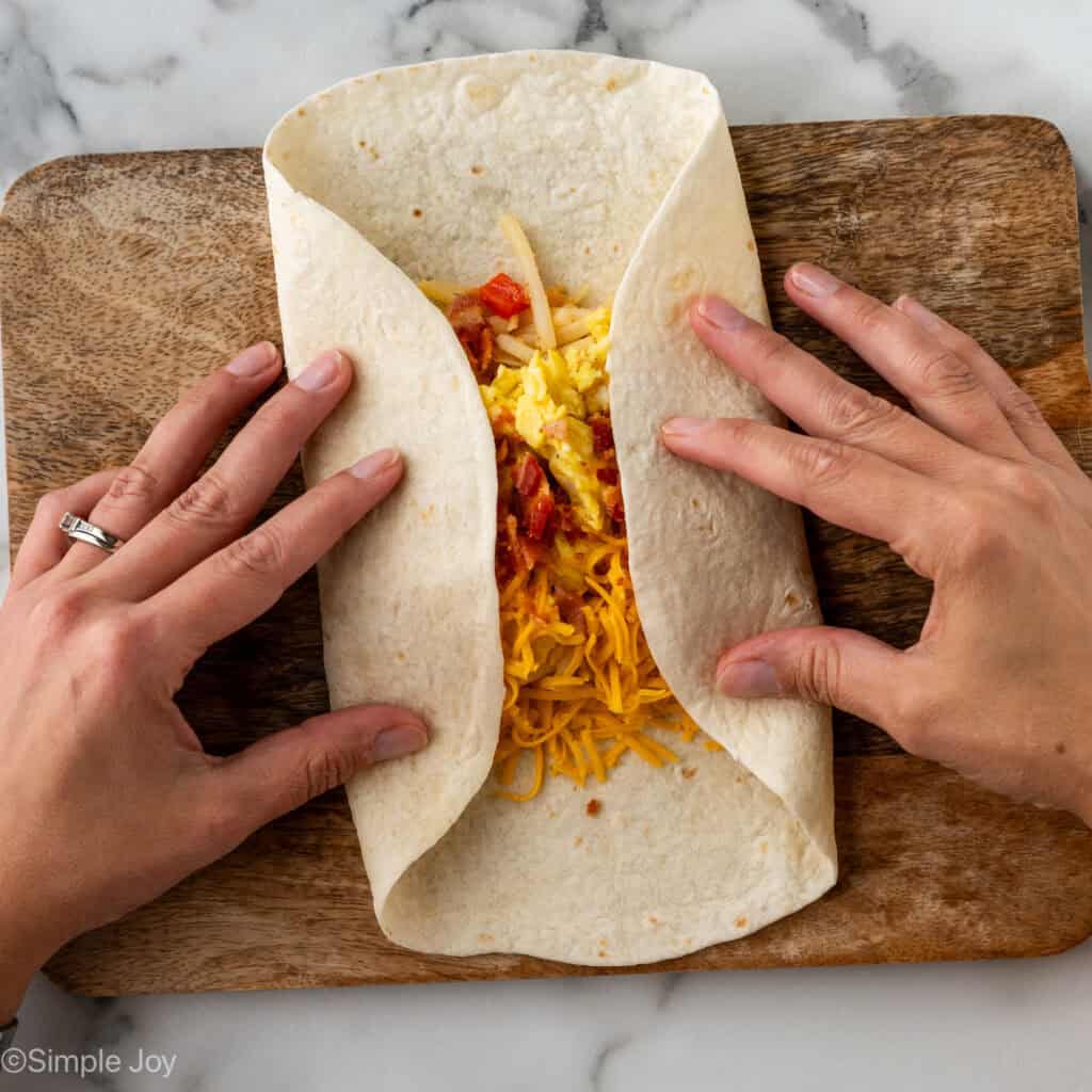 Overhead of woman's hands folding tortilla topped with Breakfast Burrito ingredients to make a breakfast burrito