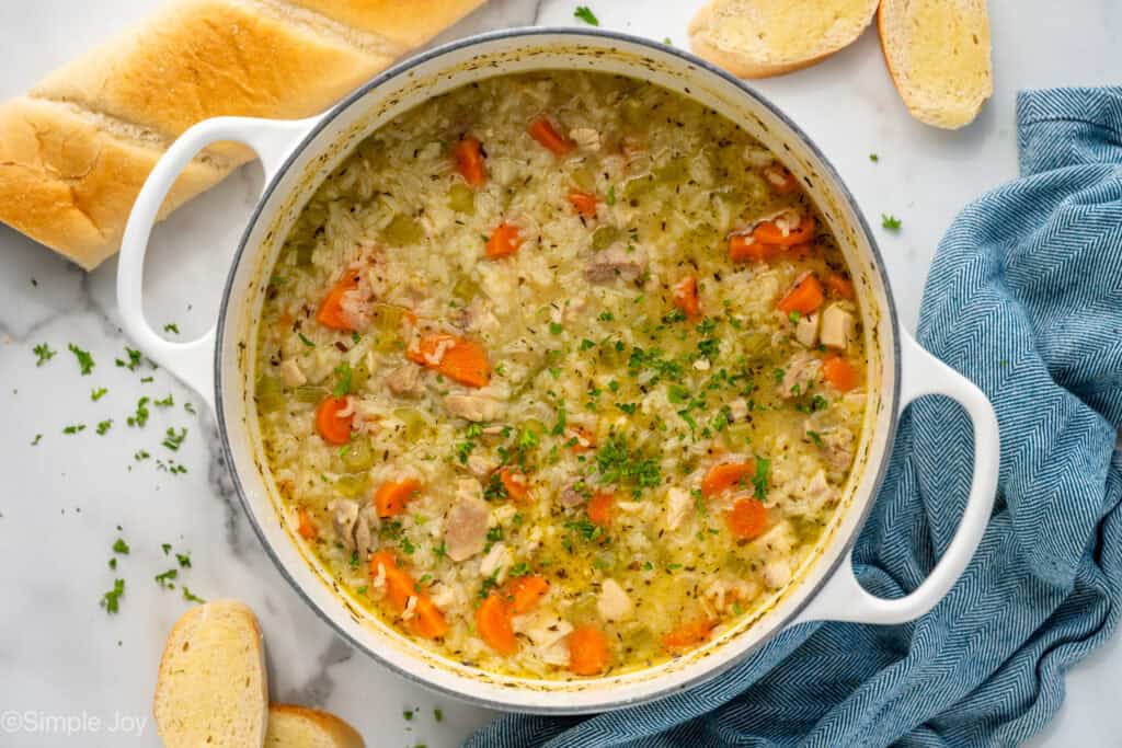 Overhead of large soup pot of chicken and rice soup garnished with fresh parsley, sliced baguette sitting beside