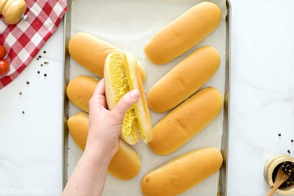 Overhead of person holding bun with mayo and mustard inside, baking sheet of hot dog buns below to make chili dog recipe.