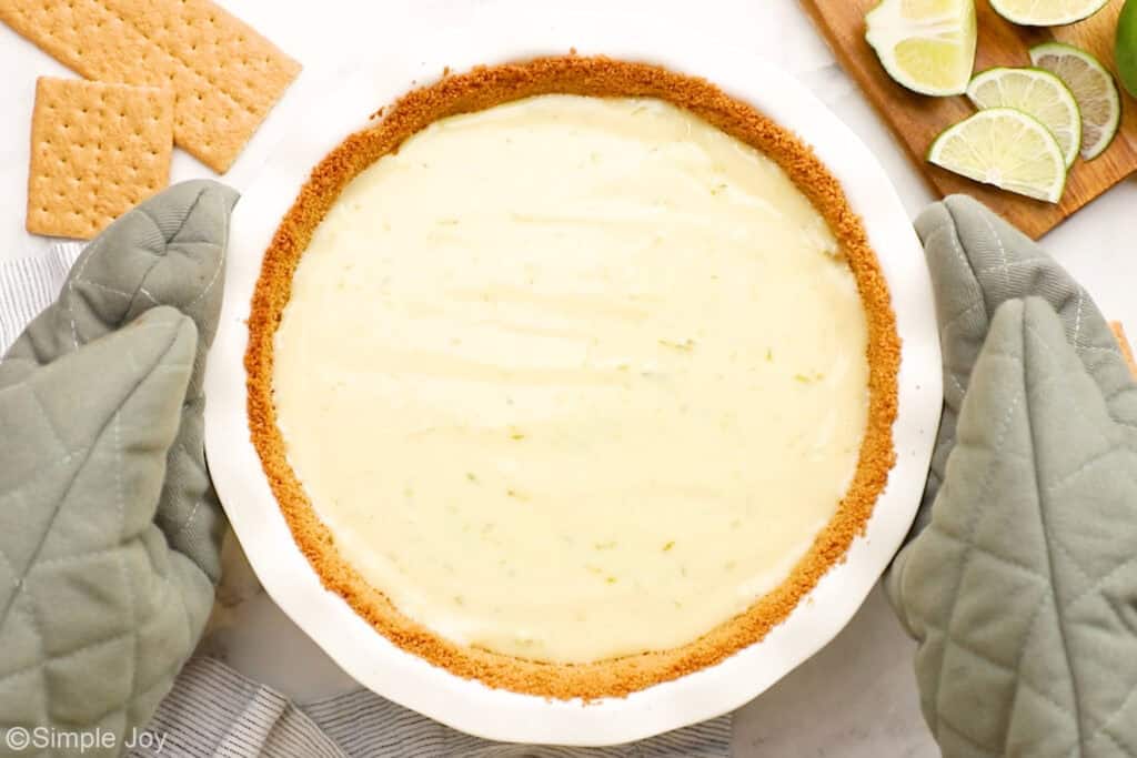 Overhead of oven mitts holding key lime pie, graham crackers and sliced limes sitting beside