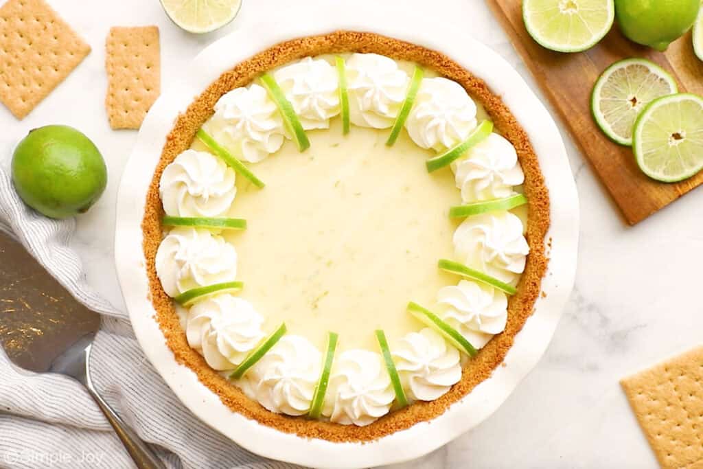 Overhead of key lime pie garnished with whipped cream and lime slices. Graham crackers, fresh limes, and serving spatula sitting beside.
