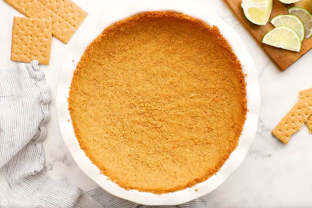 Overhead of graham cracker crust in pie plate to make key lime pie recipe. Graham crackers and sliced limes sitting beside
