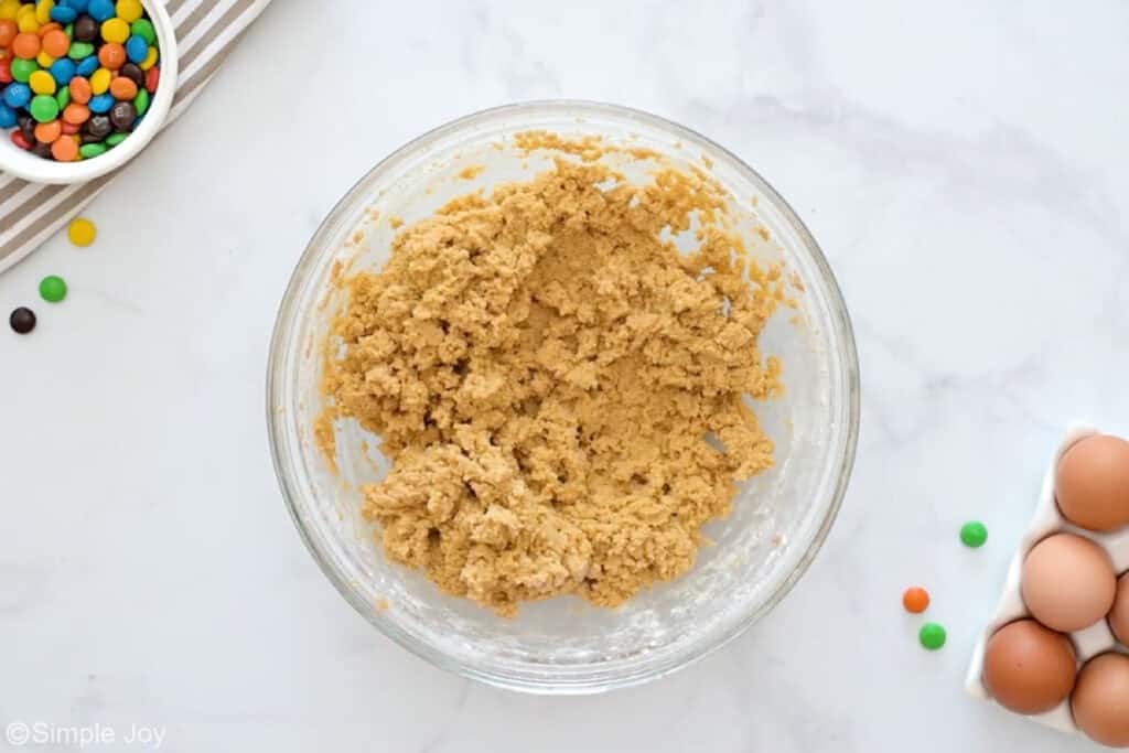 Overhead of glass bowl of m&m bar ingredients with bowl of m&ms and eggs sitting beside