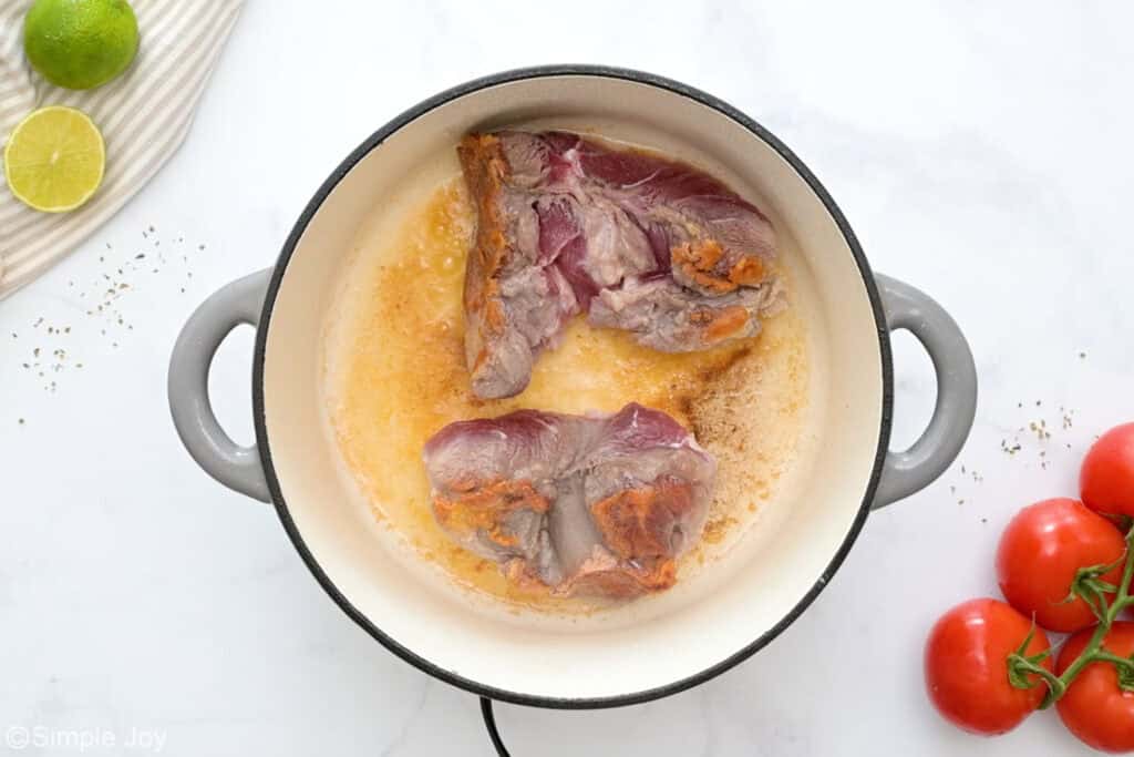 Overhead of stock pot of slow cooker carnitas ingredients, tomatoes and limes sitting beside