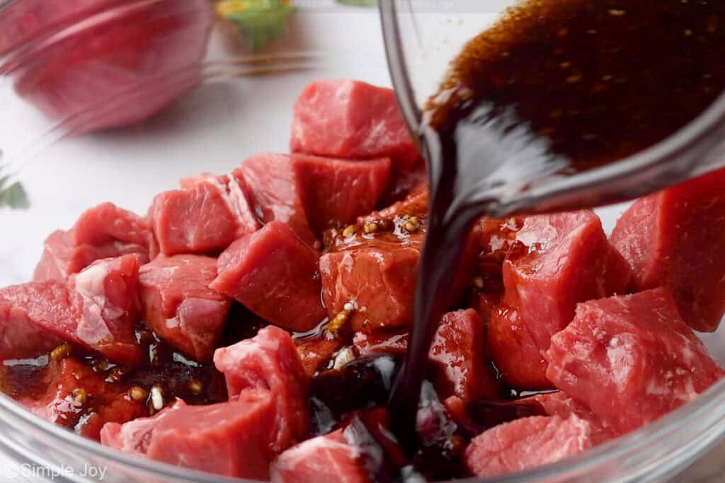 Glass measuring cup of steak marinade ingredients pouring into bowl of steak to make steak kabobs