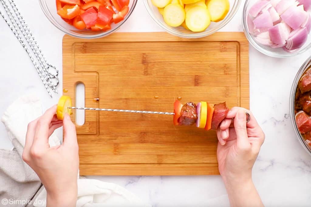 Overhead of person's hands putting vegetables and steak onto a skewer to make steak kabobs. Bowls of steak kabob ingredients sitting next to wooden cutting board