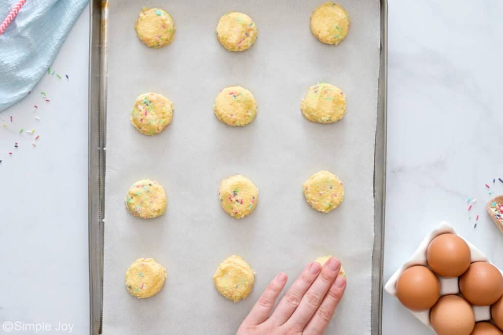 Overhead of sugar cookie dough on a parchment lined baking sheet with person's hand pressing dough ball, eggs and sprinkles sitting beside