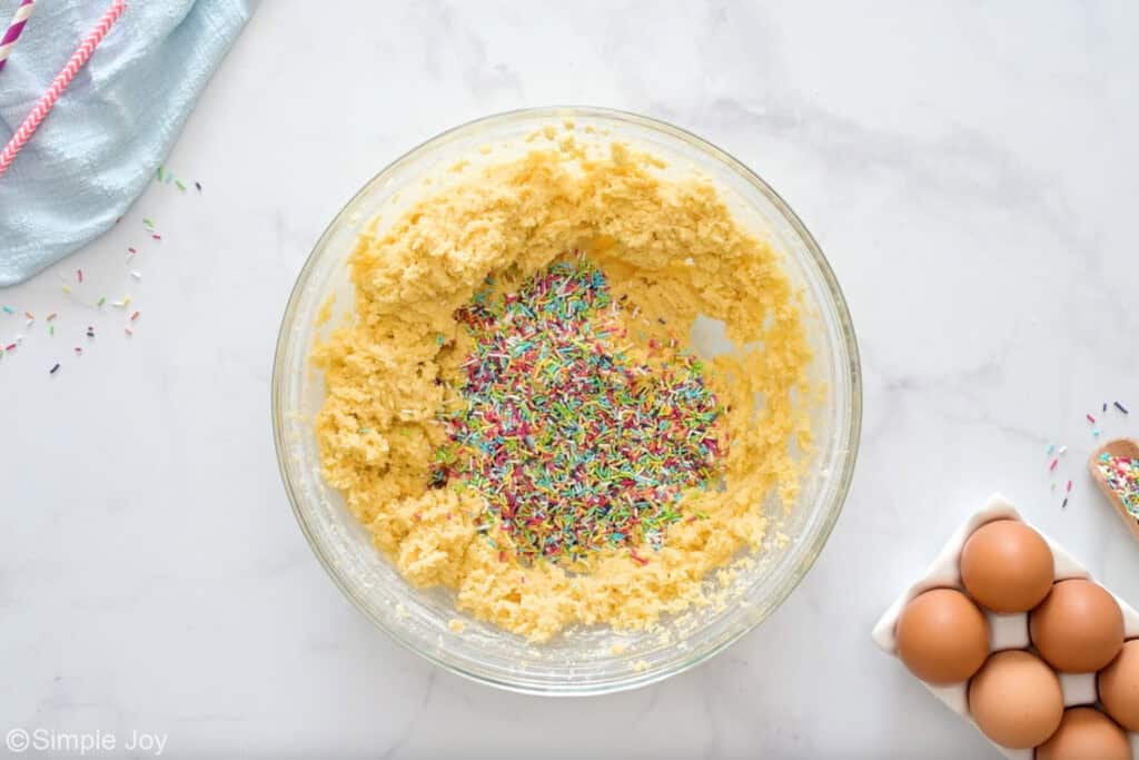 Overhead of glass bowl of sugar cookie dough ingredients with sprinkles, eggs and sprinkles sitting beside