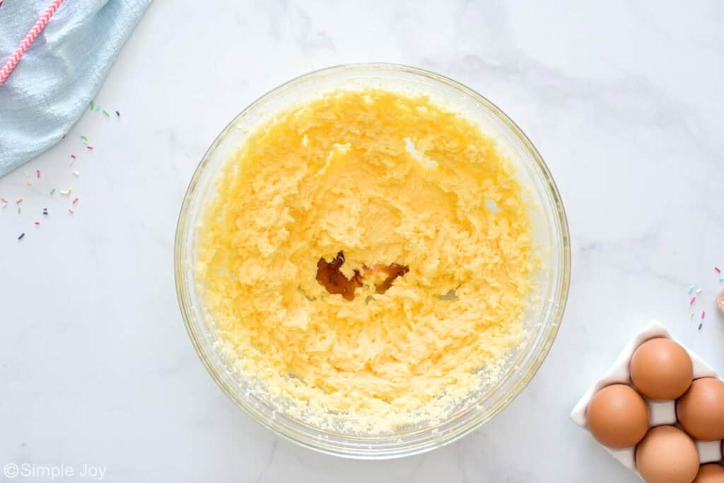 Overhead of glass bowl of sugar cookie ingredients, eggs and sprinkles sitting beside.