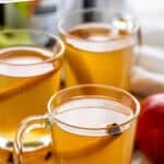 Pinterest graphic for crockpot apple cider. Says "slow cooker apple cider recipe simplejoy.com" Image shows three mugs of crockpot apple cider with cinnamon sticks inside and beside