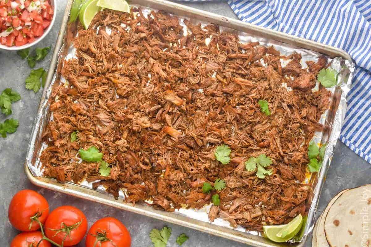 baking sheet full of the pulled meat from a carnitas recipe topped with fresh cilantro and two limes