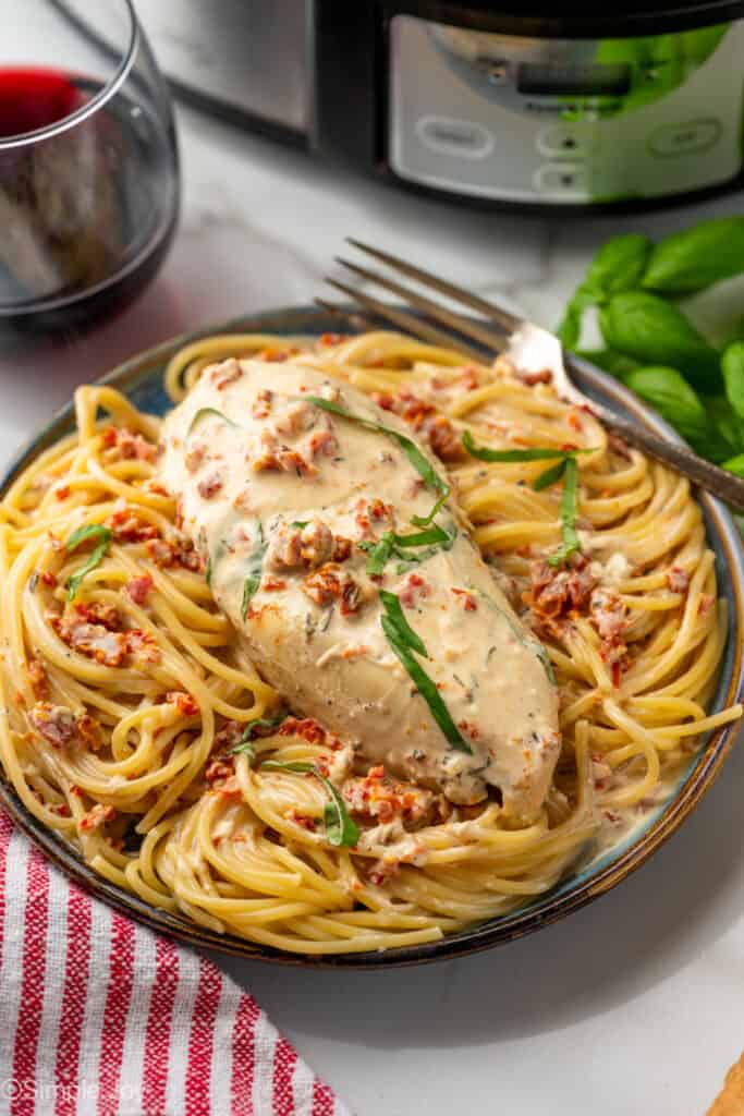 Plate of pasta topped with marry me chicken with crockpot and glass of red wine sitting in background
