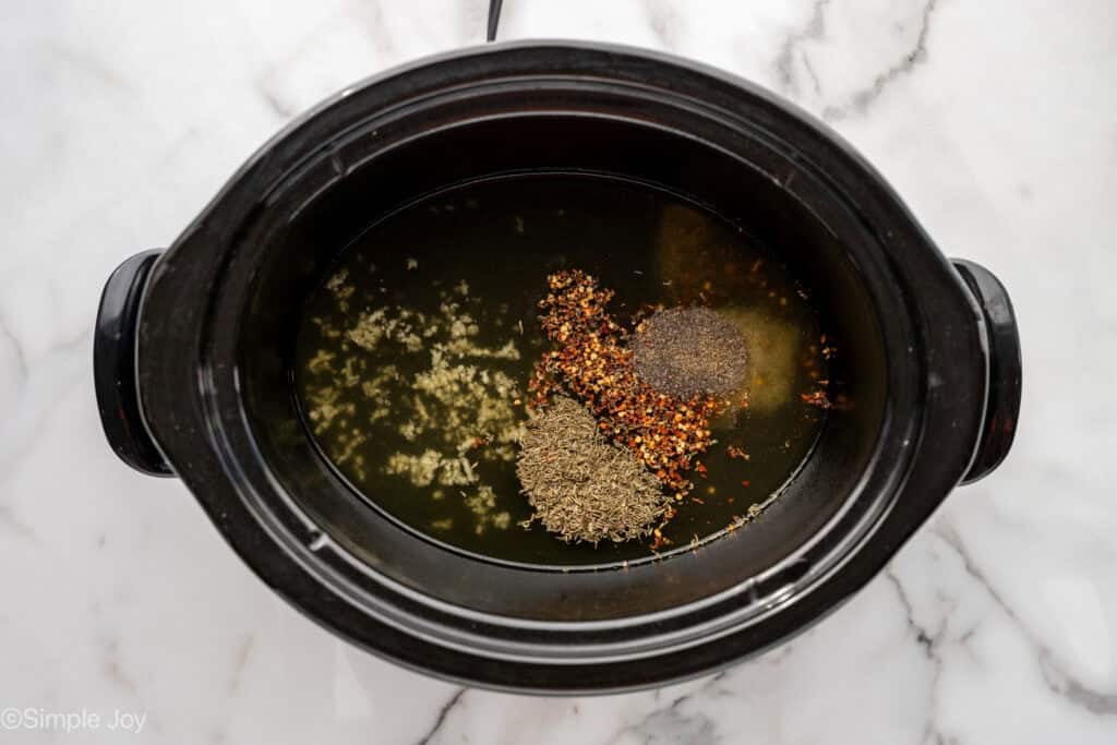 overhead of a slow cooker with chicken broth, garlic, thyme, salt, crushed red pepper flakes, and black pepper