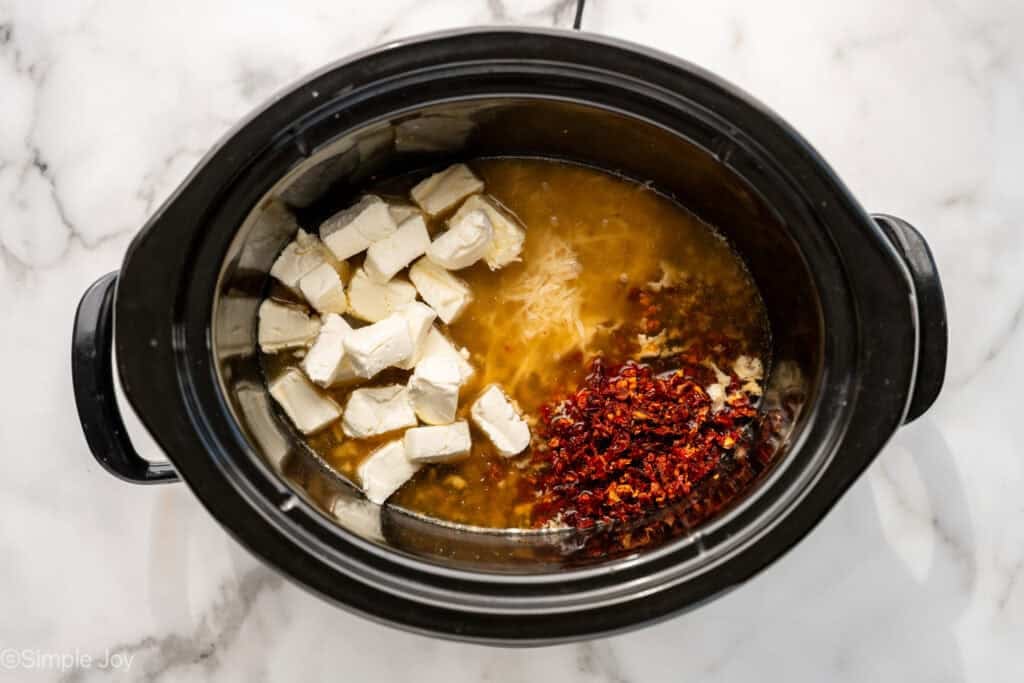 overhead of slow cooker with cream cheese cut up, diced sun-dried tomatoes, and grated parmesan cheese