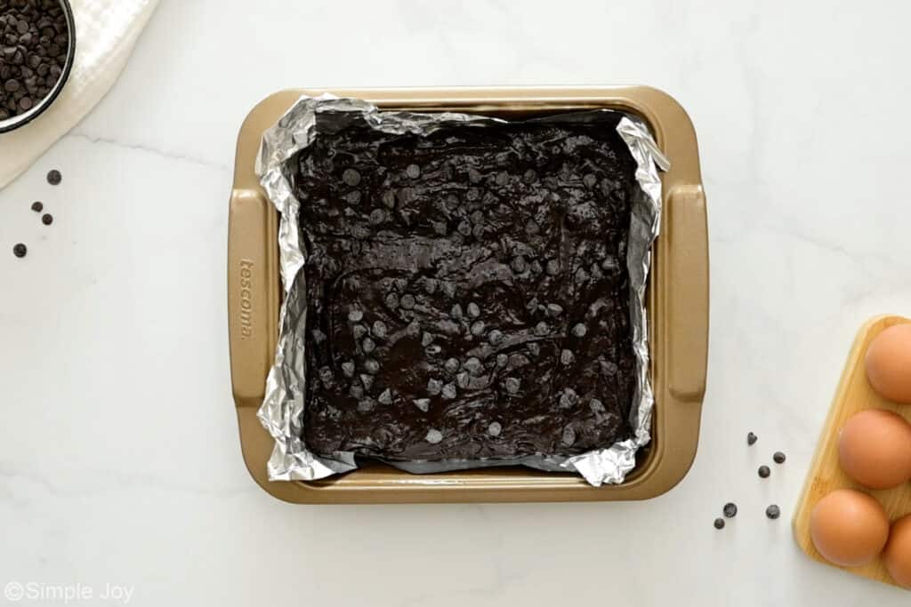 Overhead of foil-lined baking pan of death by chocolate brownies batter, eggs and chocolate chips sitting beside