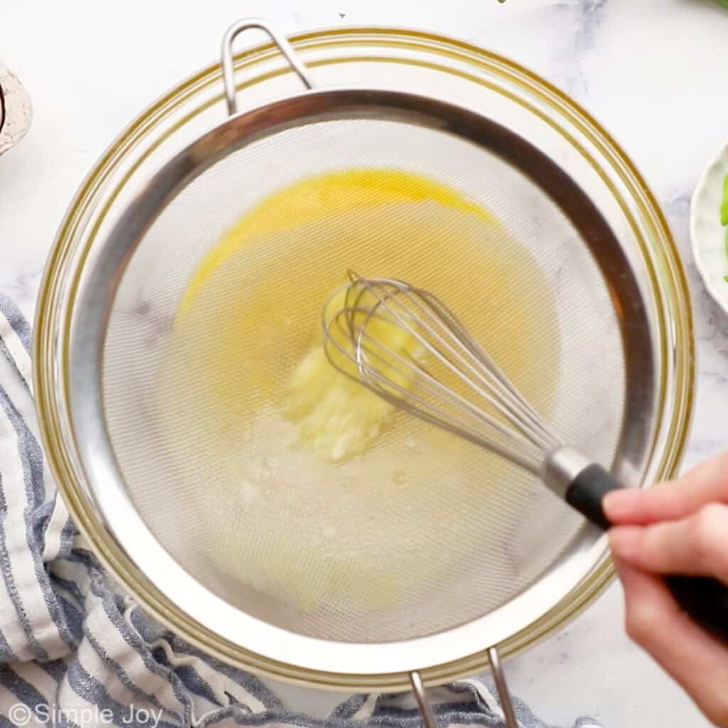 Overhead of eggs in metal strainer over bowl with person's hand holding whisk to make egg drop soup