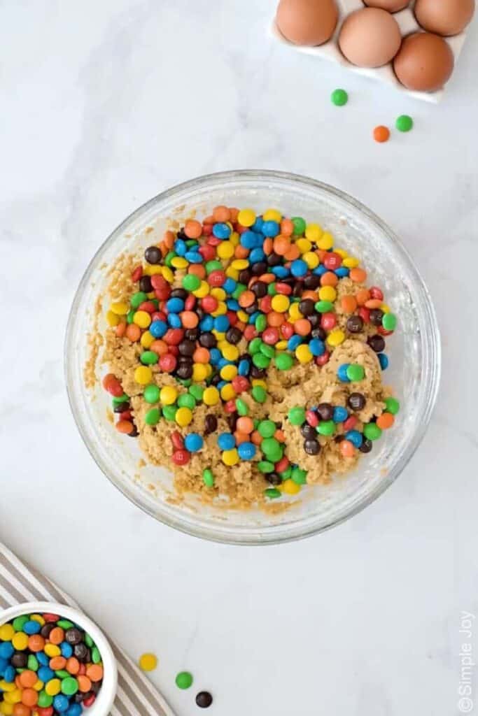 Overhead of bowl of m&m bars ingredients, bowl of m&m candy and eggs sitting beside