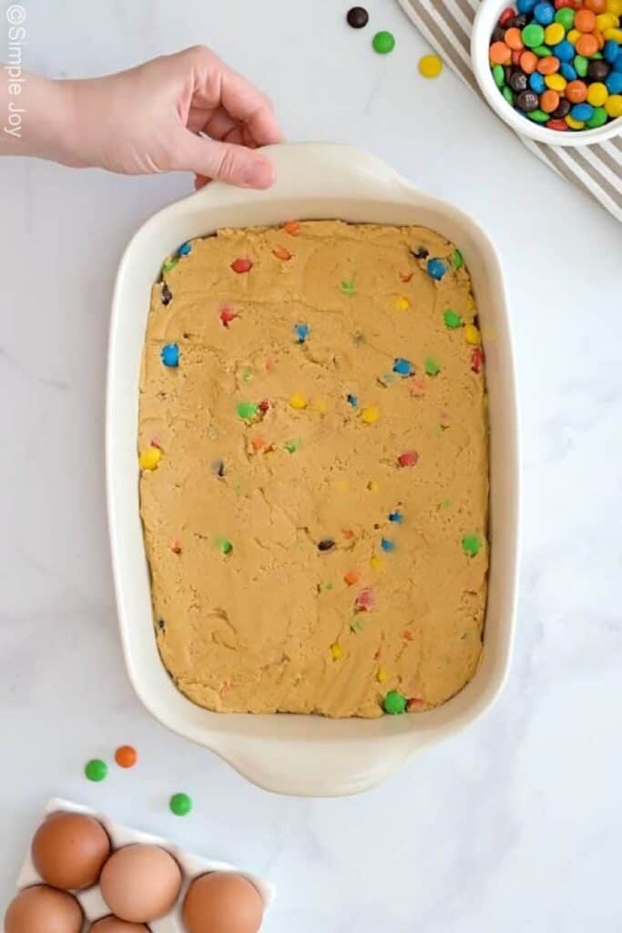 Overhead of person's hand next to m&m cookie bar dough in a baking dish to make m&m bars. Bowl of m&m candies and eggs sitting beside
