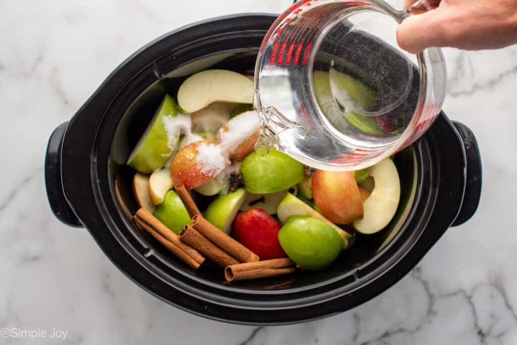 Person's hand pouring measuring cup of water into slow cooker of crockpot apple cider ingredients