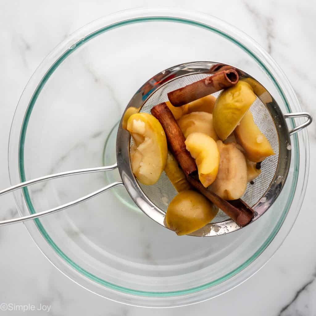 Overhead of cooked apples and cinnamon sticks in a mesh strainer over a glass bowl to make crockpot apple cider