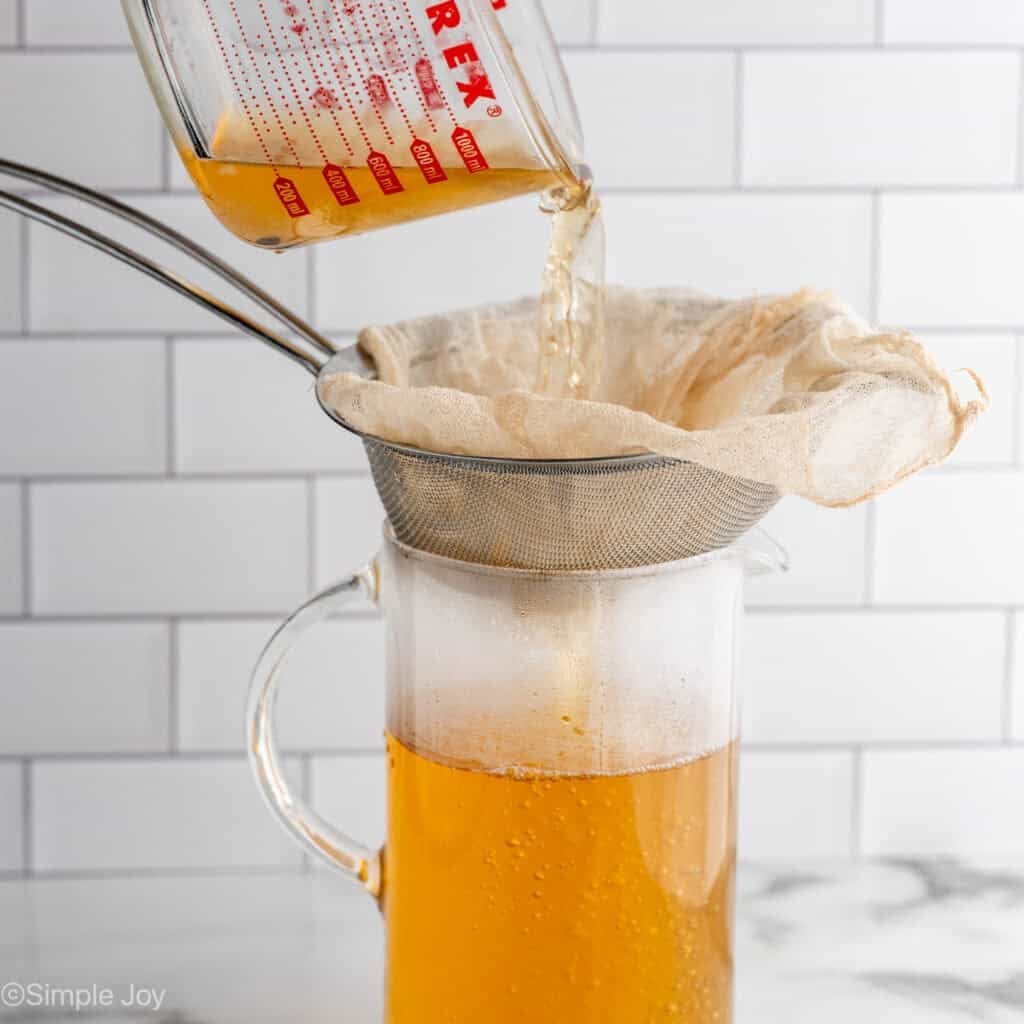 Glass measuring cup of crockpot apple cider pouring over mesh strainer into a glass pitcher