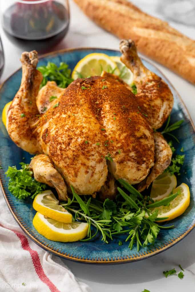 Instant Pot Whole Chicken on a platter surrounded by lemon slices and fresh herbs. Glass of red wine and baguette sitting in background