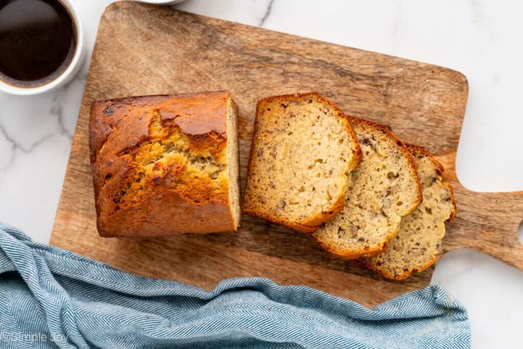 Overhead of slices and loaf of banana bread on a wooden board with cup of black coffee sitting beside