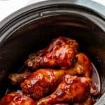 Pinterest graphic for crockpot chicken drumsticks. Says "slow cooker chicken drumsticks simplejoy.com" Image shows a crockpot of chicken drumsticks ingredients