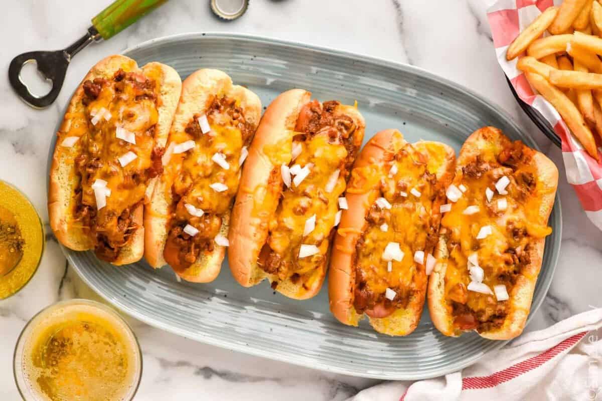 Overhead of plate of Chili Dogs topped with diced onion. Side of french fries and two glasses of beer sitting beside