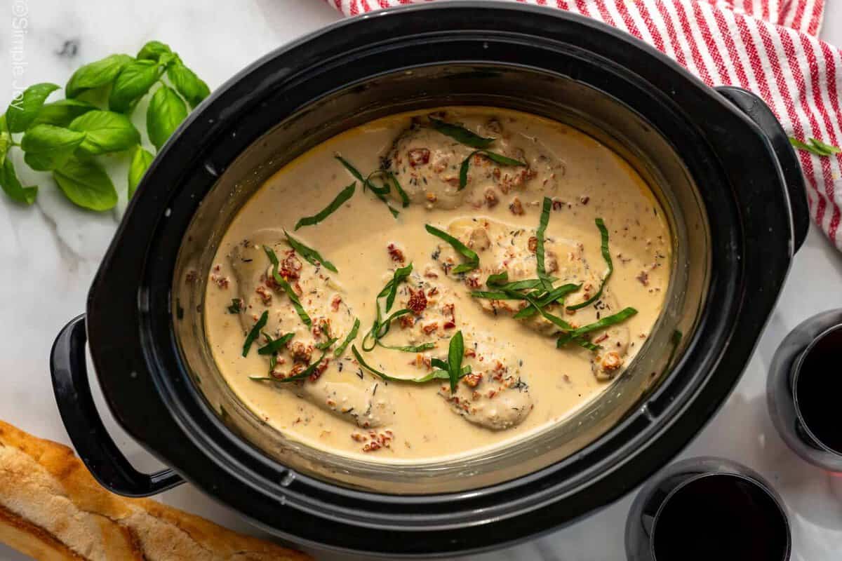 Overhead of slow cooker of Marry Me Chicken garnished with fresh basil leaves. Fresh basil leaves, baguette, and two glasses of red wine surrounding