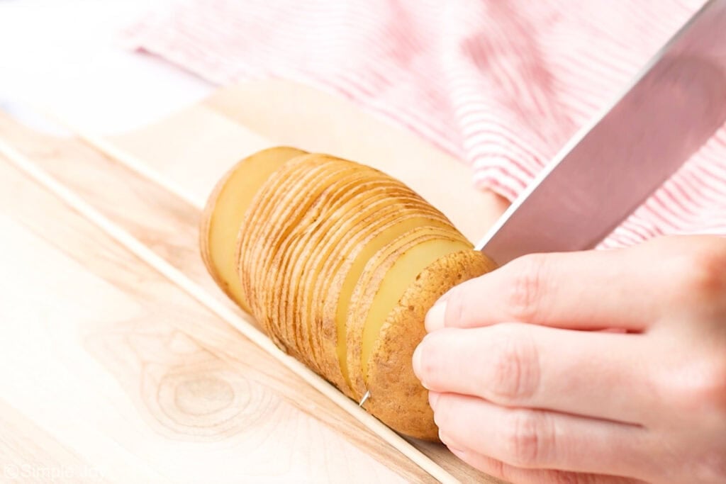 Person holding potato with large knife slices into potato to make hasselback potatoes