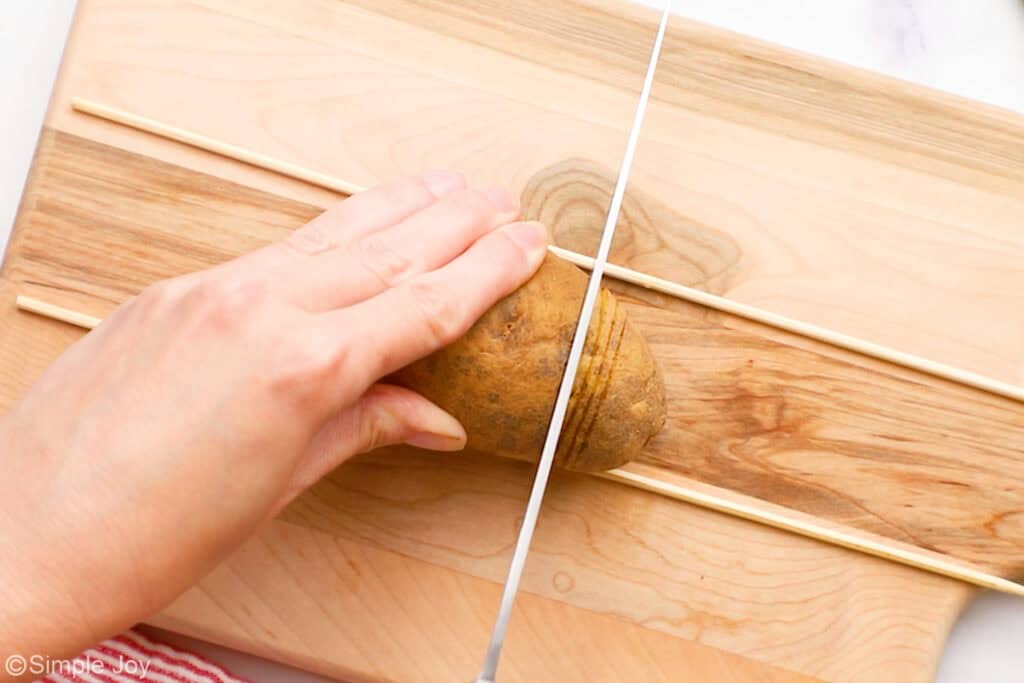 Overhead of person's hand holding potato on a wooden cutting board while large knife slices into potato to make hasselback potatoes