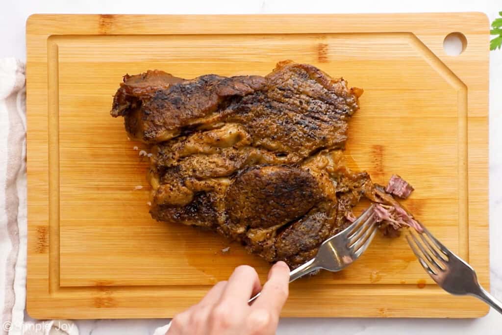 Overhead of slow cooked pot roast on a wooden cutting board with person shredding the roast with two forks