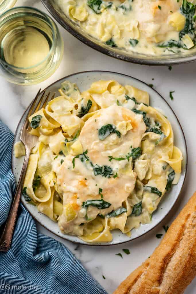 Overhead of plate of spinach artichoke chicken over pasta with fork. Glass of white wine and skillet of spinach artichoke chicken sitting behind.