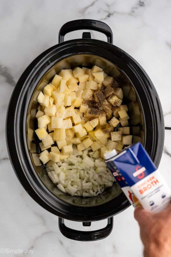 Overhead of a crockpot with diced potatoes, onions, and seasonings with person's hand pouring chicken broth into slow cooker