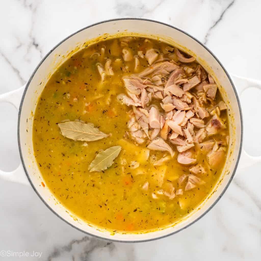 overhead of a dutch oven full of broth with vegetables, diced chicken, and two bay leaves