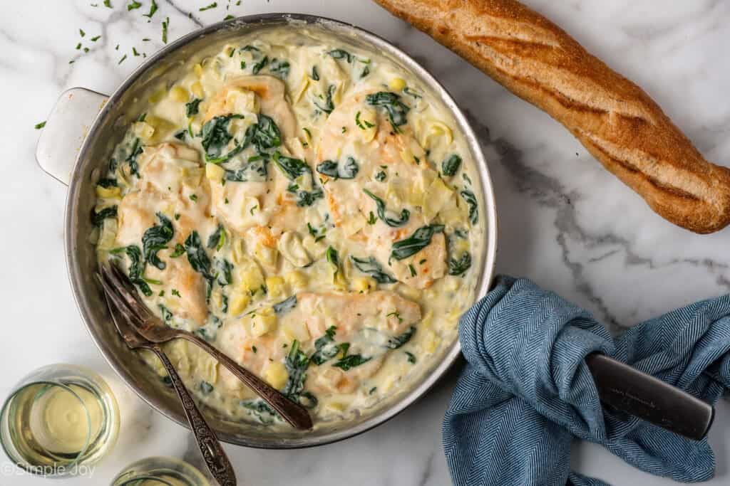 Overhead of skillet of spinach artichoke chicken with two forks. Baguette and two glasses of white wine sitting beside
